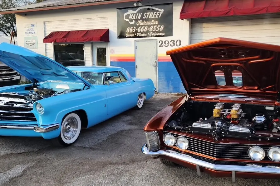 Two classic cars with open hoods outside the shop